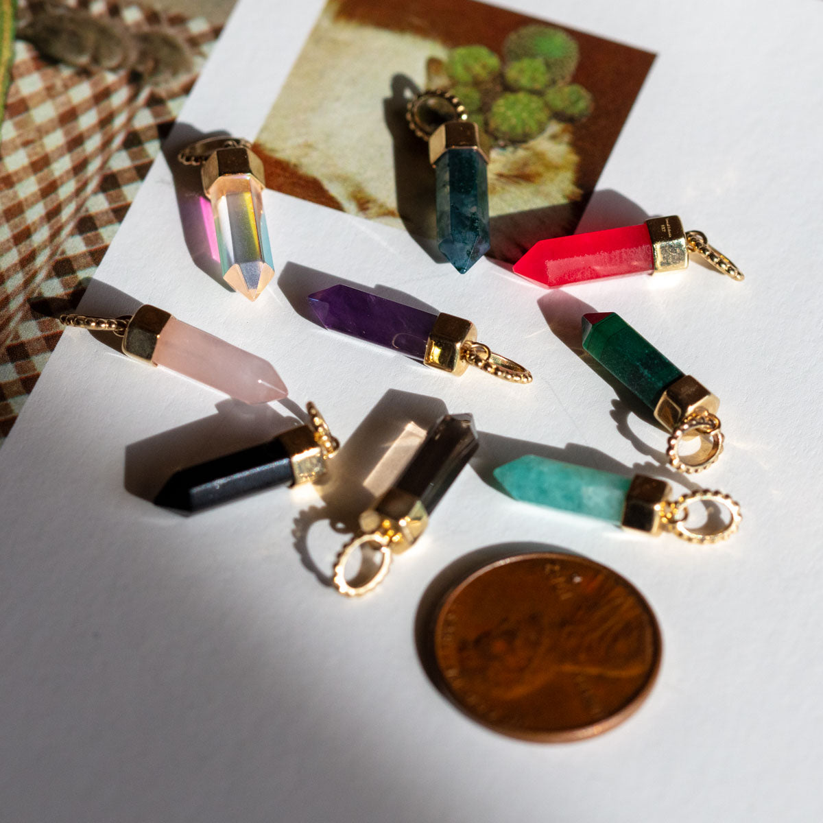 A Solid Gold Crystal Charm for Manifestation is shown with gold-capped natural crystals arranged on a white surface, a penny for scale. A softly blurred cactus photo in the background adds charm and manifesting energy to the display.