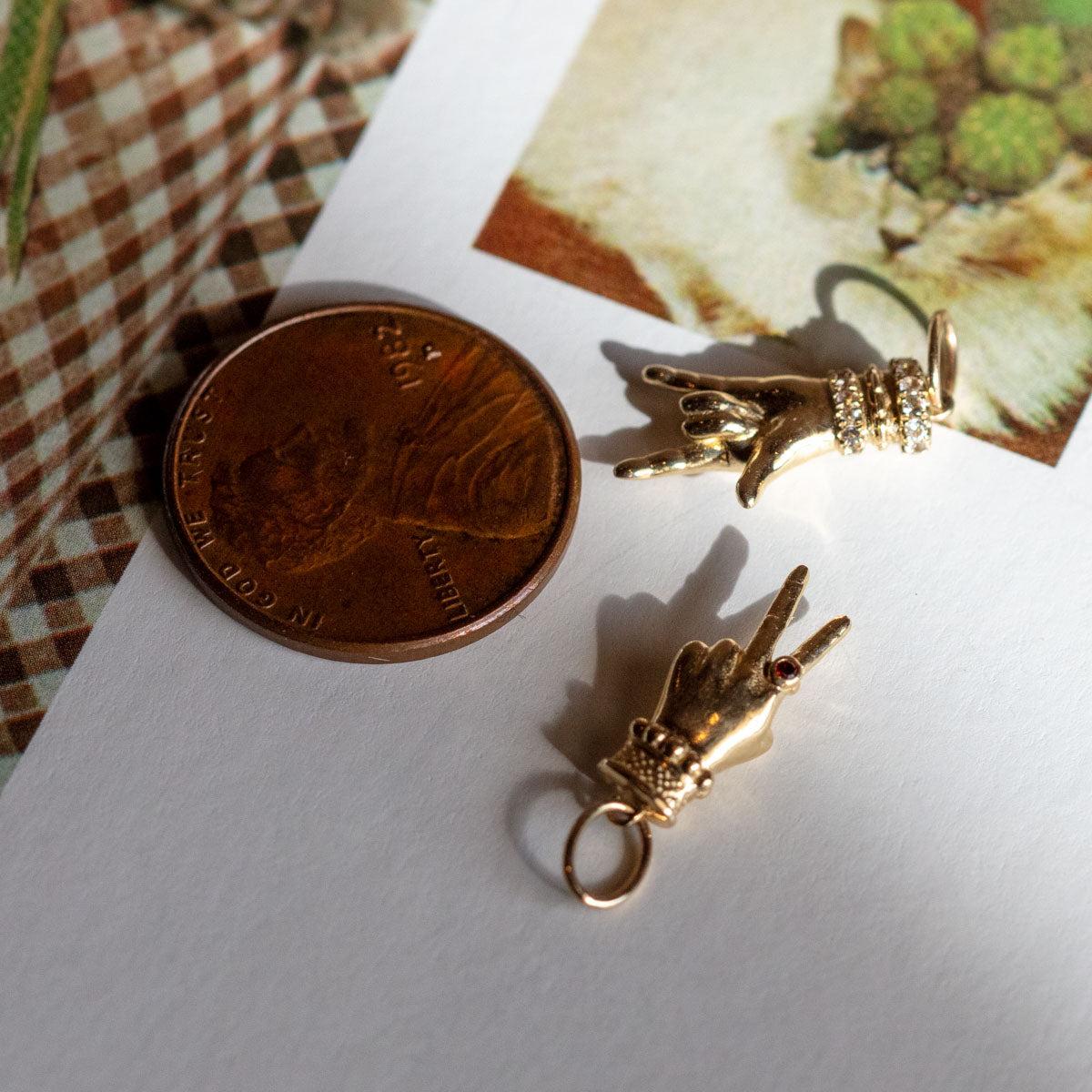 A U.S. penny is shown next to two Solid Gold Body Language Charms making the ASL I love you sign, all resting on a cat photo—an exclusive from Local Eclectic.