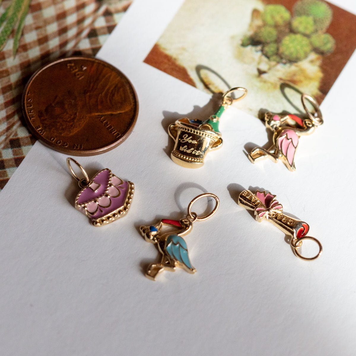 Five Solid Gold Celebration Charms are displayed on a white surface next to a penny for scale; their colorful enamel designs have gold edges, with a lion postcard and plaid fabric partially visible in the background.