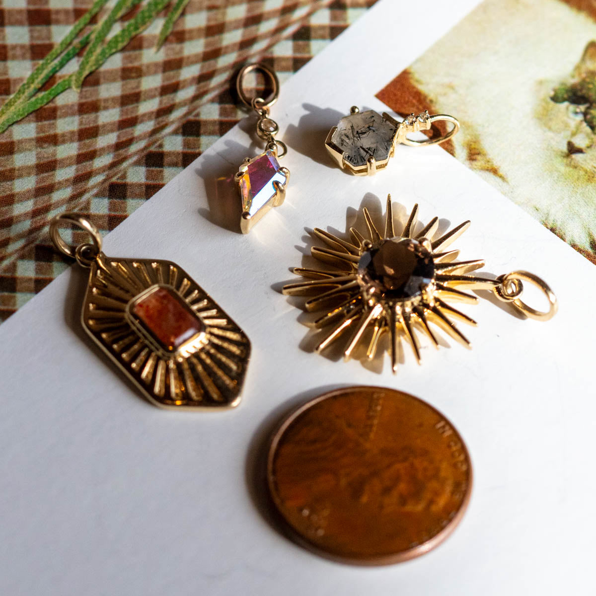 Four gold pendants, including a Solid Gold Amulet Charm, are arranged on a white surface next to a copper coin. The pendants display sunburst and rectangular designs, set against a plaid fabric background.
