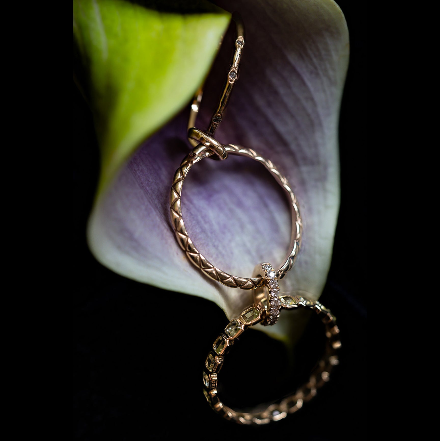 The Solid Gold Delicate Woven Stacking Ring, adorned with intricate patterns and small gemstones, rests on soft purple and green flower petals, standing out vividly against the dark background.