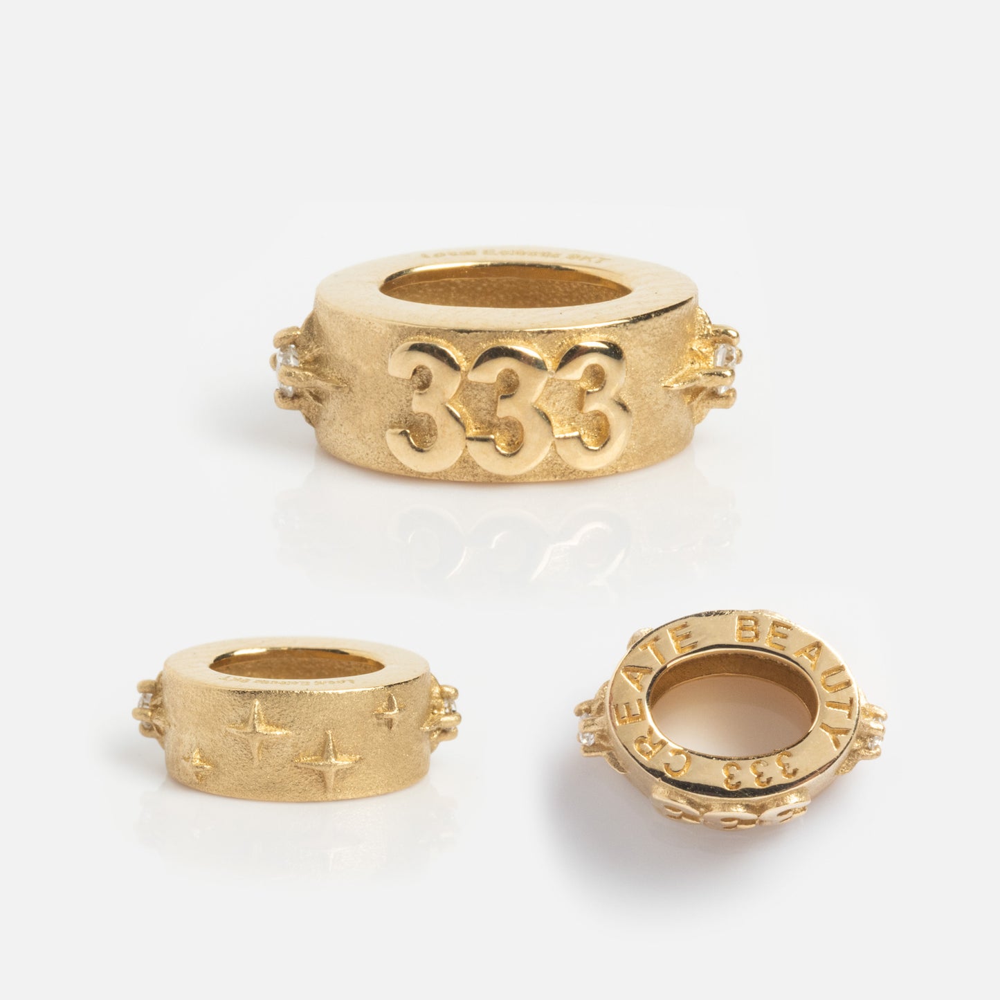 The Solid Gold Angel Number Spacer Charm set includes three matte gold barrel-shaped beads: one marked with 333, one engraved with stars, and another inscribed CREATE BEAUTY around the edge.