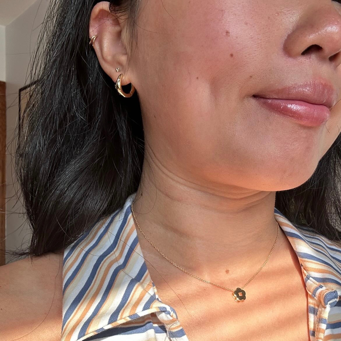 A woman with long dark hair wears the Star Crossed Celestial Hoop Earrings in gold vermeil, a small gold stud, and a gold flower pendant necklace. She is dressed in a striped shirt, bathed in natural sunlight.