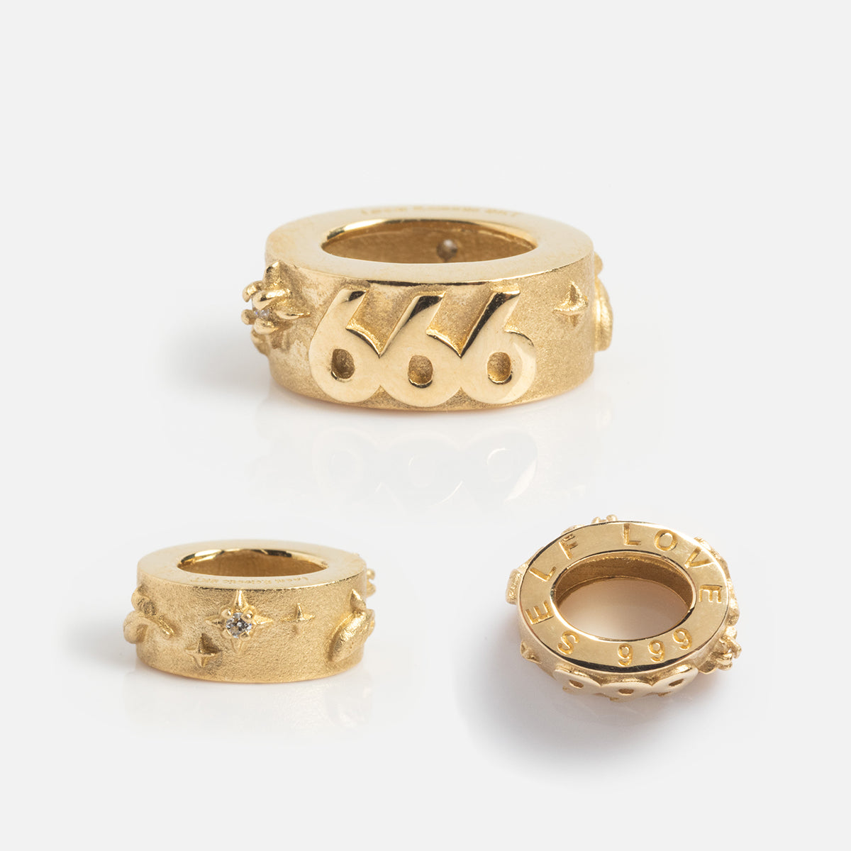 Three gold rings on a white background: a Local Eclectic Exclusive with 666, one with the Solid Gold Angel Number Spacer Charm featuring angel numbers plus star and diamond details, and a third displaying FEEL LOVE and 999.