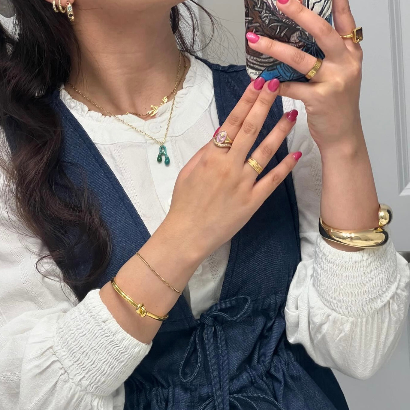 A person wearing a white blouse and denim dress holds a phone, showcasing pink manicured nails, gold rings, bracelets, earrings, wavy hair, and the Bubble Letter Necklace—a 14k gold plated necklace with bubble letter charms.