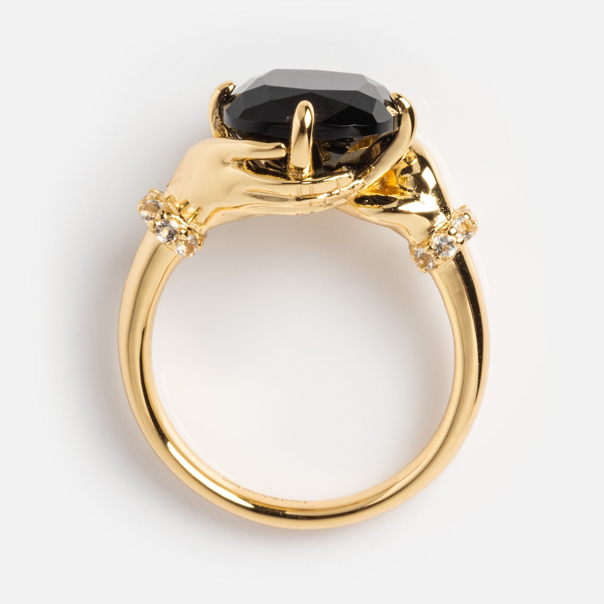 The Past Life Black Spinel Statement Ring features two sculpted hands holding a large faceted black spinel, with small diamonds on the band near each hand—a striking vintage-inspired piece.