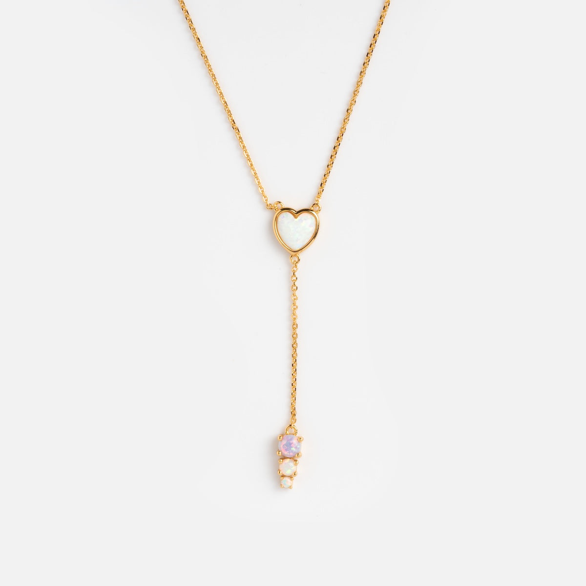 The Opal Heart Lariat features a heart-shaped opal pendant centered on a gold vermeil lariat necklace, with a vertical drop chain ending in three small round gemstones, shown against a plain white background.