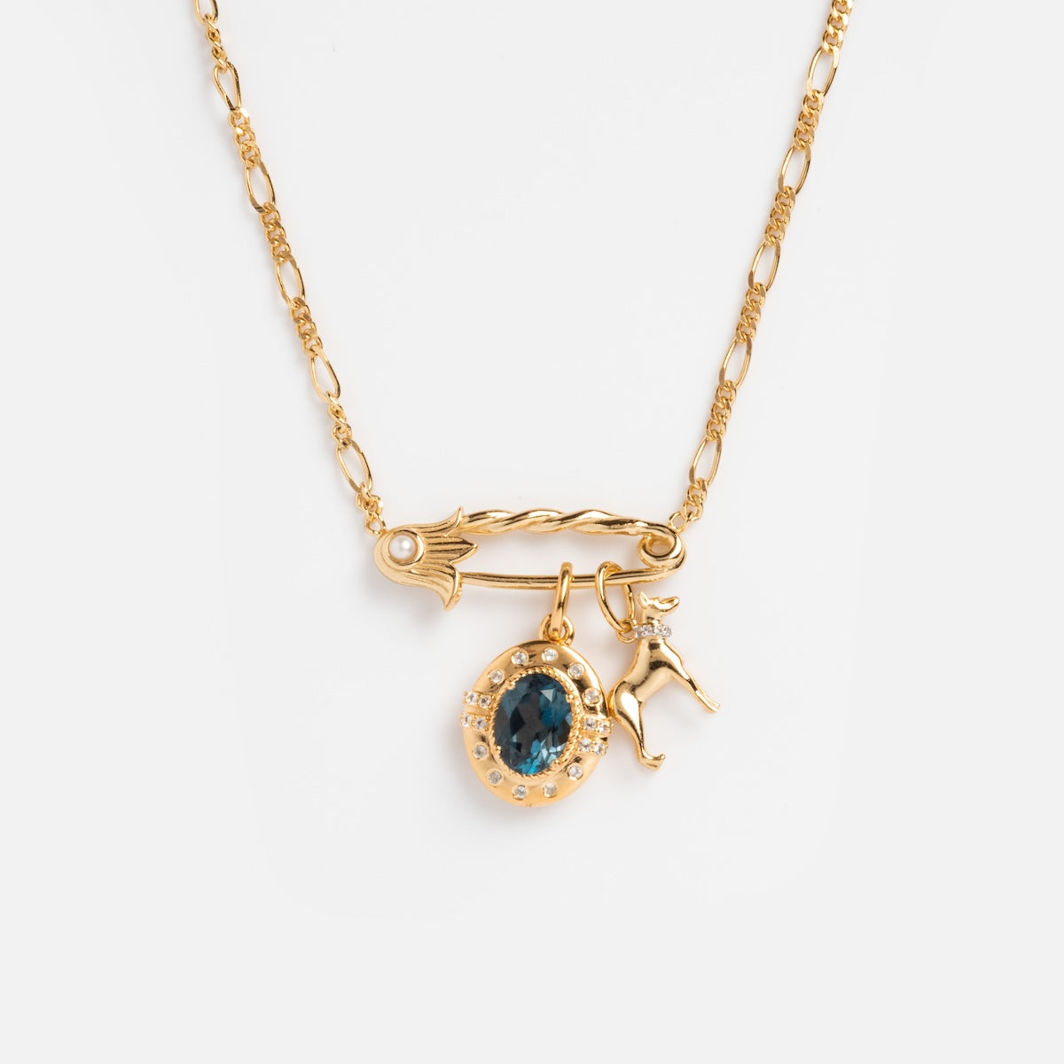 The Sonnet Safety Pin Charm Holder Necklace features a twisted gold pin clasp inspired by art deco, with an oval blue gemstone charm surrounded by clear stones and a small gold deer figurine, shown on a plain white background.