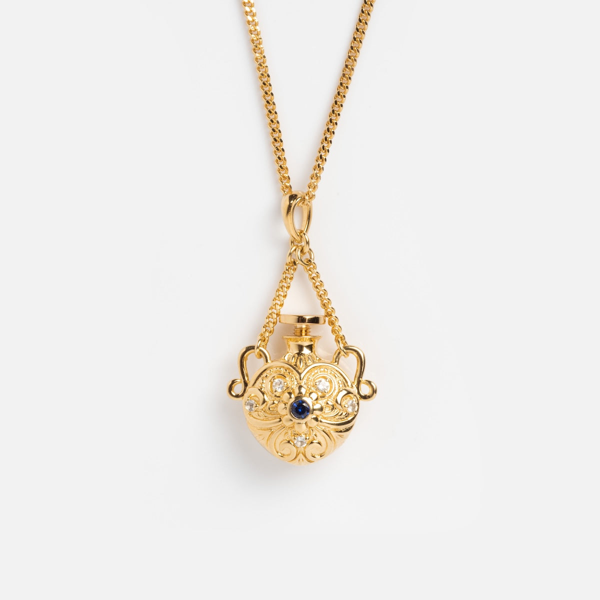 The Juliet Vial Pendant Necklace features an ornate gold flask-shaped pendant with intricate scrollwork and a central blue sapphire, hanging from a delicate gold chain—a stunning piece of vintage-inspired jewelry.