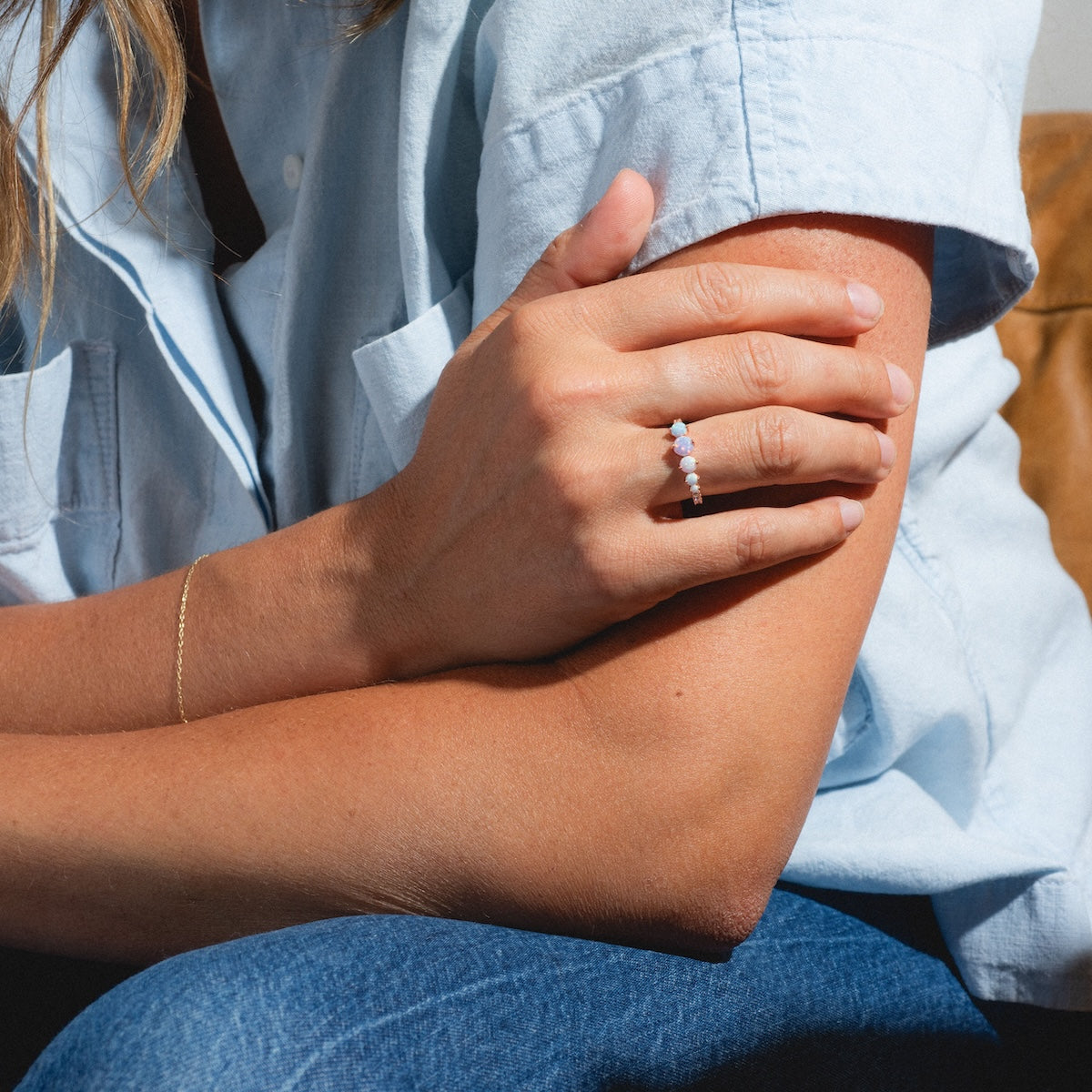 A person in a light blue short-sleeve shirt and jeans showcases their delicate gold bracelet and the Pastel Opal Shooting Star Ring on their finger. The image highlights their hands and upper body, with the ring featuring five opals.