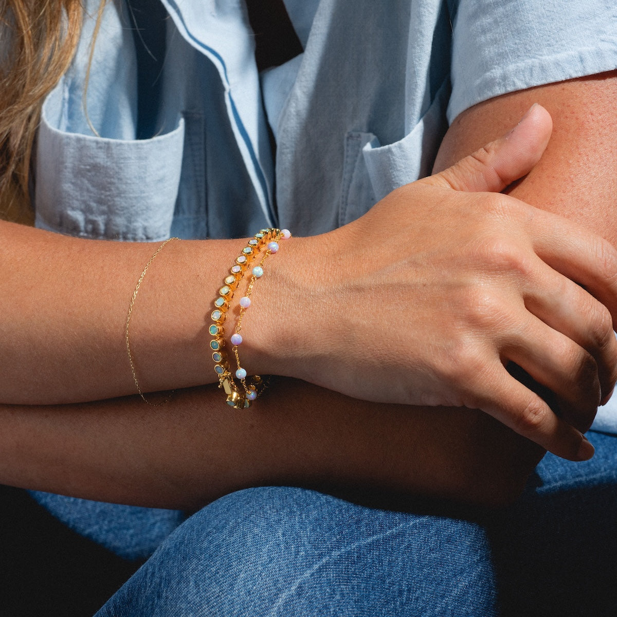 A person in a light blue short-sleeve shirt and blue jeans sits with crossed arms, showcasing the Pastel Opal Gum Drop Bracelet and a gold vermeil bracelet on their wrist. Bright lighting highlights the shimmer of the jewelry.