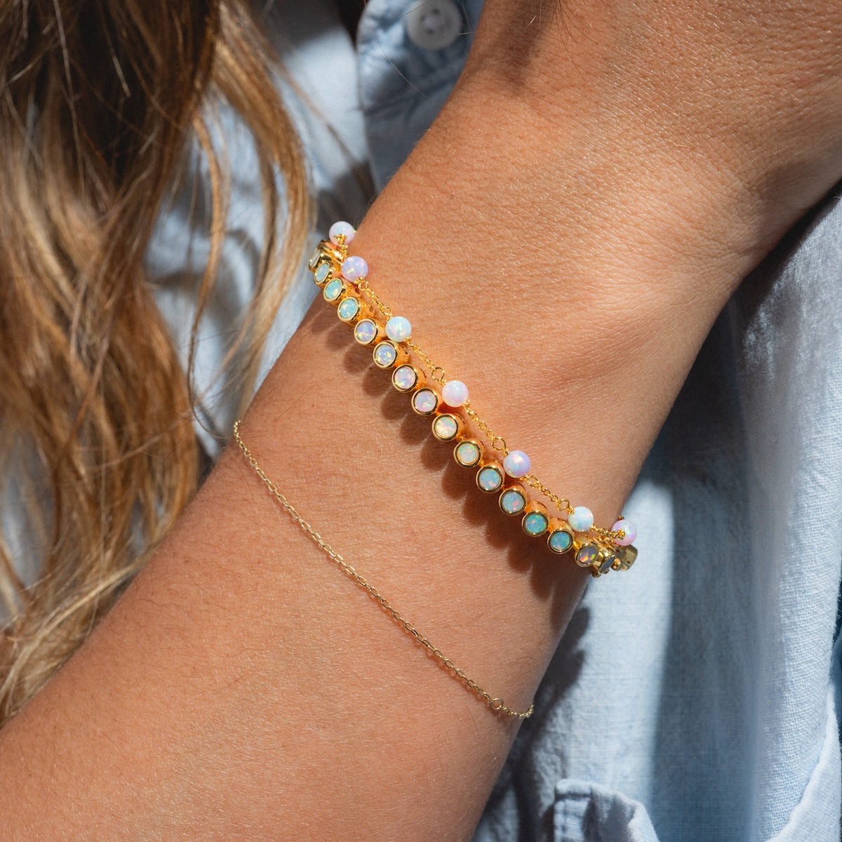 A close-up of a wrist wearing a gold vermeil chain and the Pastel Opal Ombre Tennis Bracelet, featuring pastel opals set in gold. The person sports a light blue shirt, with brown hair partially visible.