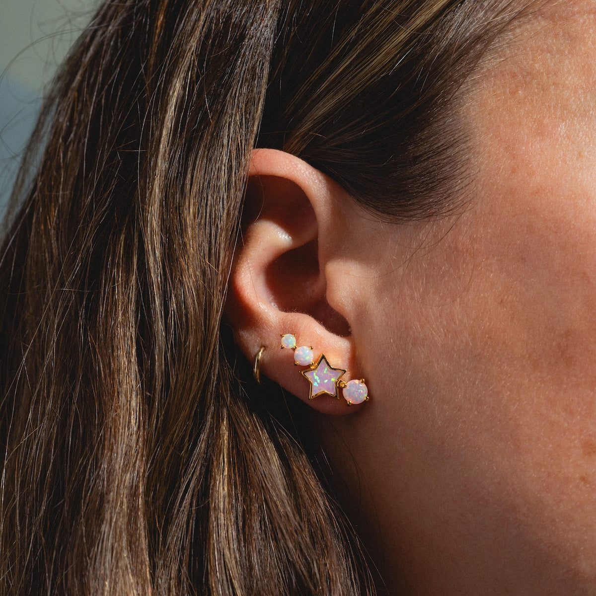 A close-up of a woman’s ear with four earrings, including the Pastel Opal Shooting Star Studs—opal celestial studs—plus a small gold hoop and a gold star earring, set against brown hair and sunlit skin.
