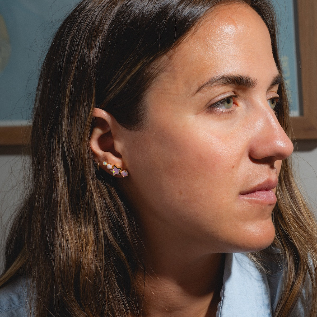 A woman with long brown hair, wearing Pastel Opal Shooting Star Studs, looks to the right in soft lighting. She has a neutral expression, wears a light blue shirt, and stands before a blurred background with a framed picture.