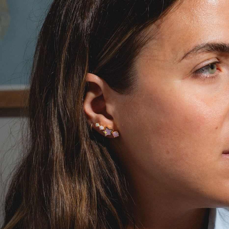 A woman with long brown hair, wearing Pastel Opal Shooting Star Studs, looks to the right in soft lighting. She has a neutral expression, wears a light blue shirt, and stands before a blurred background with a framed picture.
