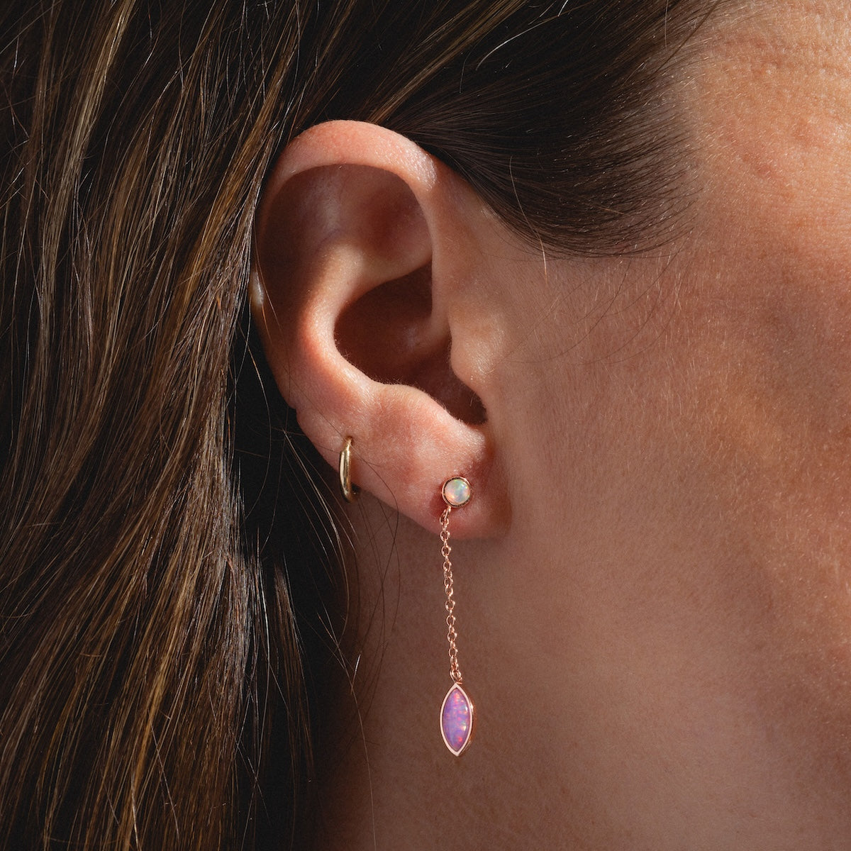 Close-up of a womans ear wearing three earrings: a gold hoop, the Rose Gold Opal Dew Drop Studs in the middle lobe, and a dangling earring with a pink gemstone at the bottom.