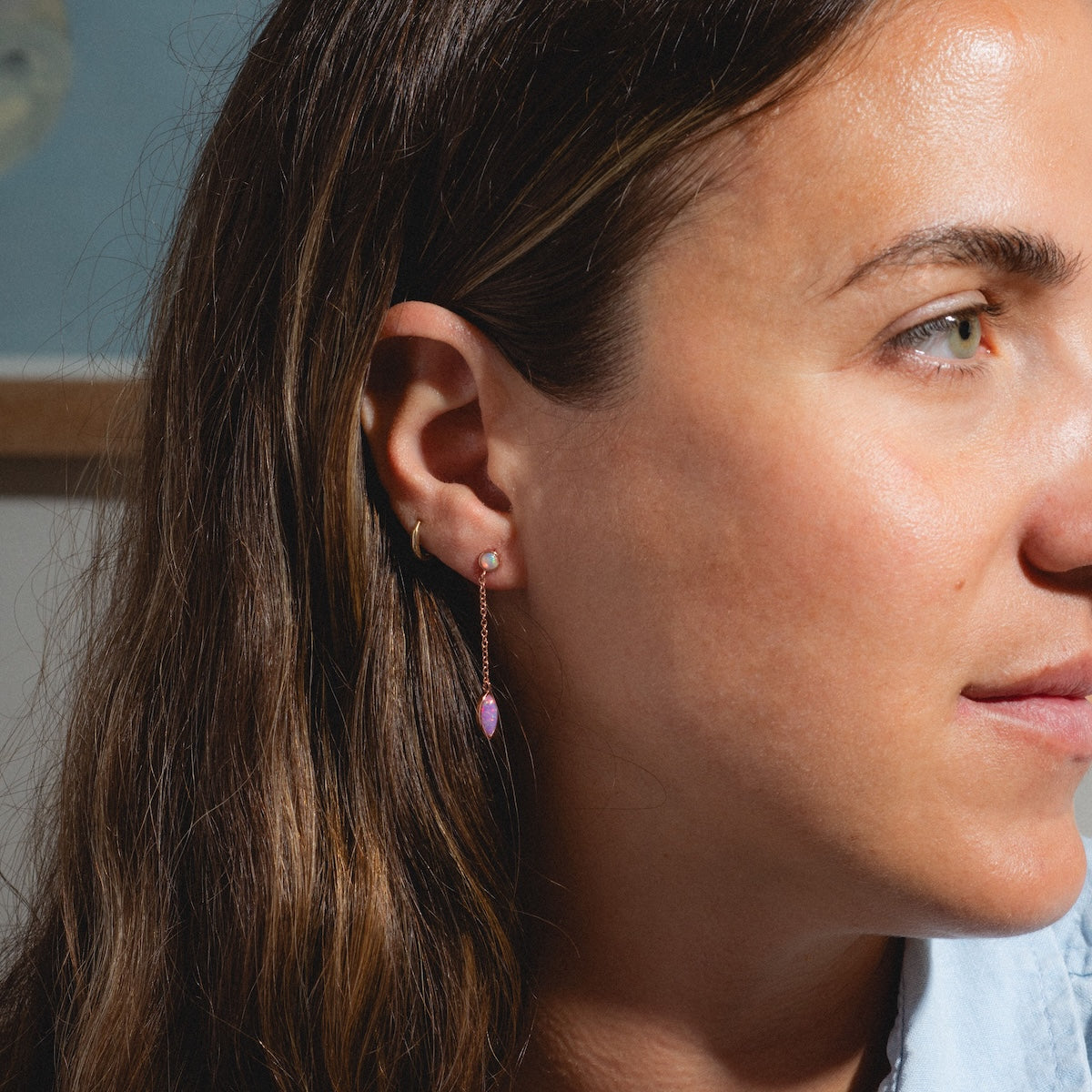 Close-up of a woman with long brown hair wearing Rose Gold Opal Dew Drop Studs—delicate teardrop-shaped pink opal earrings—as she looks slightly to the right. A blurred map is visible in the background.