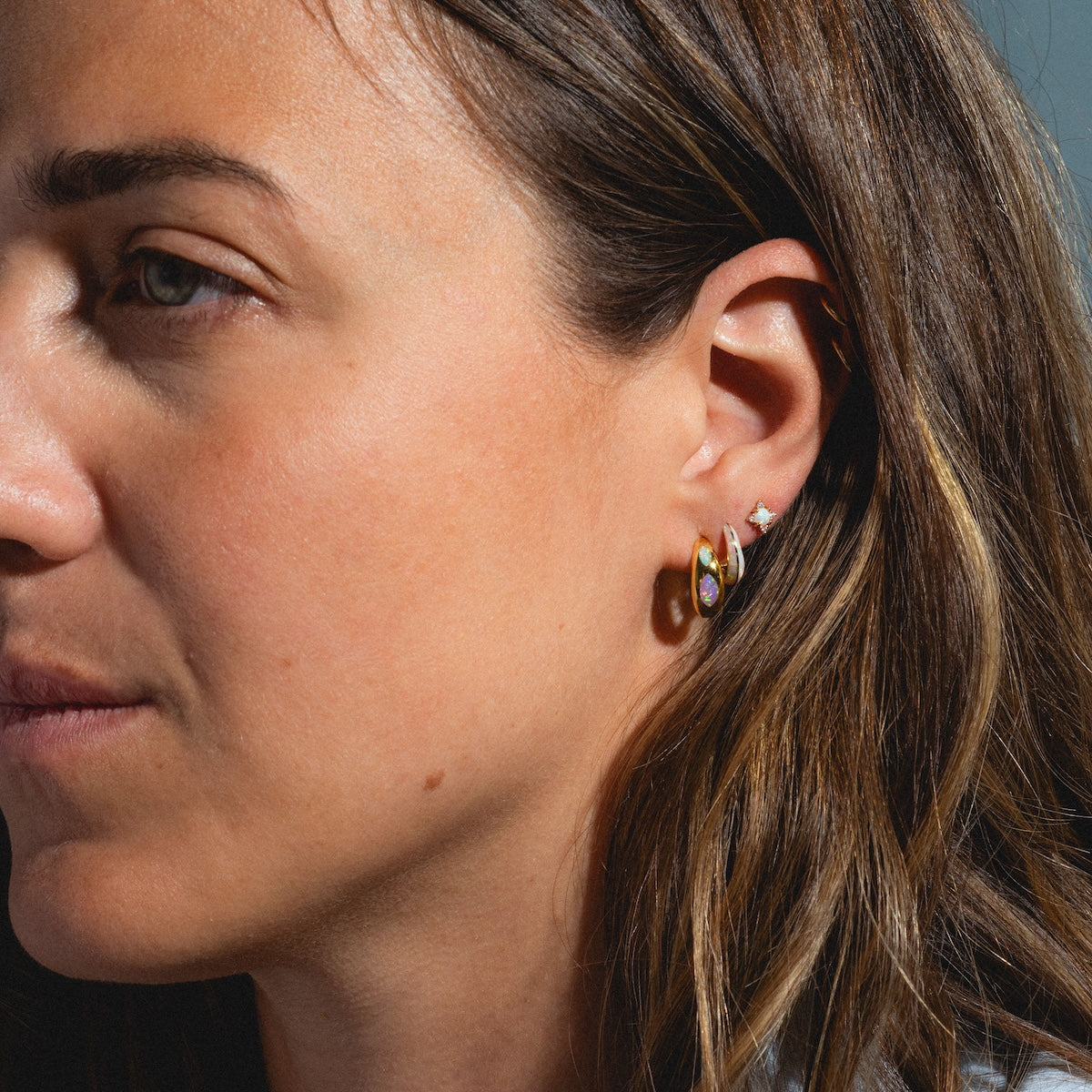 Close-up of a womans face displaying multiple earrings, including small hoops and Starlight Studs with white opals, complemented by her light brown hair and natural makeup.