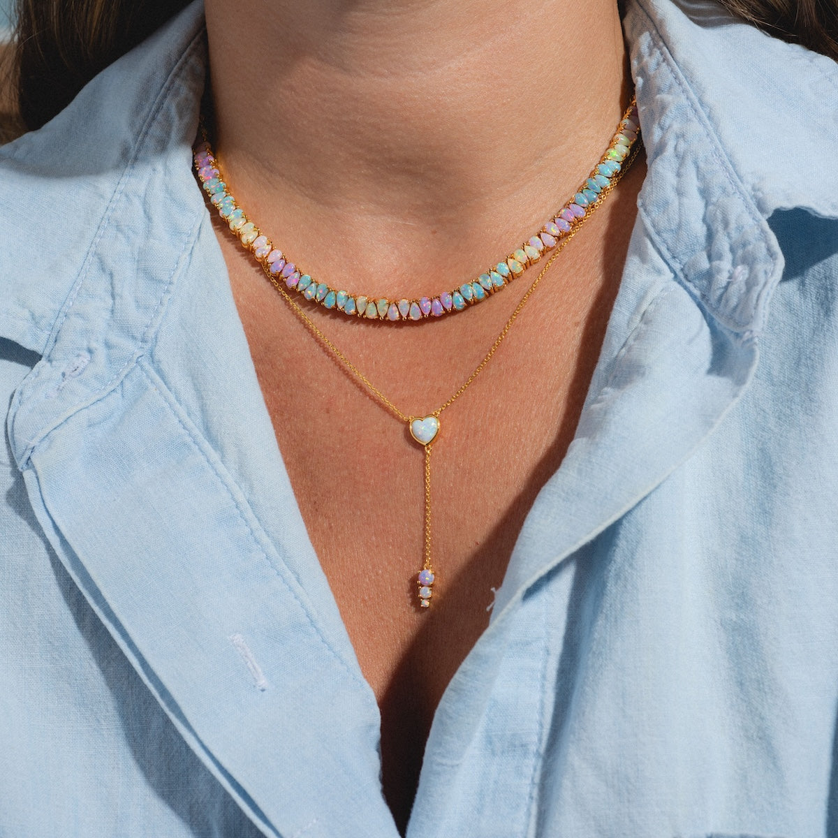 A person in a light blue, open-collar shirt wears two necklaces: a colorful beaded choker and the Opal Heart Lariat, a gold vermeil chain featuring a small heart-shaped opal pendant with a dangling jewel.