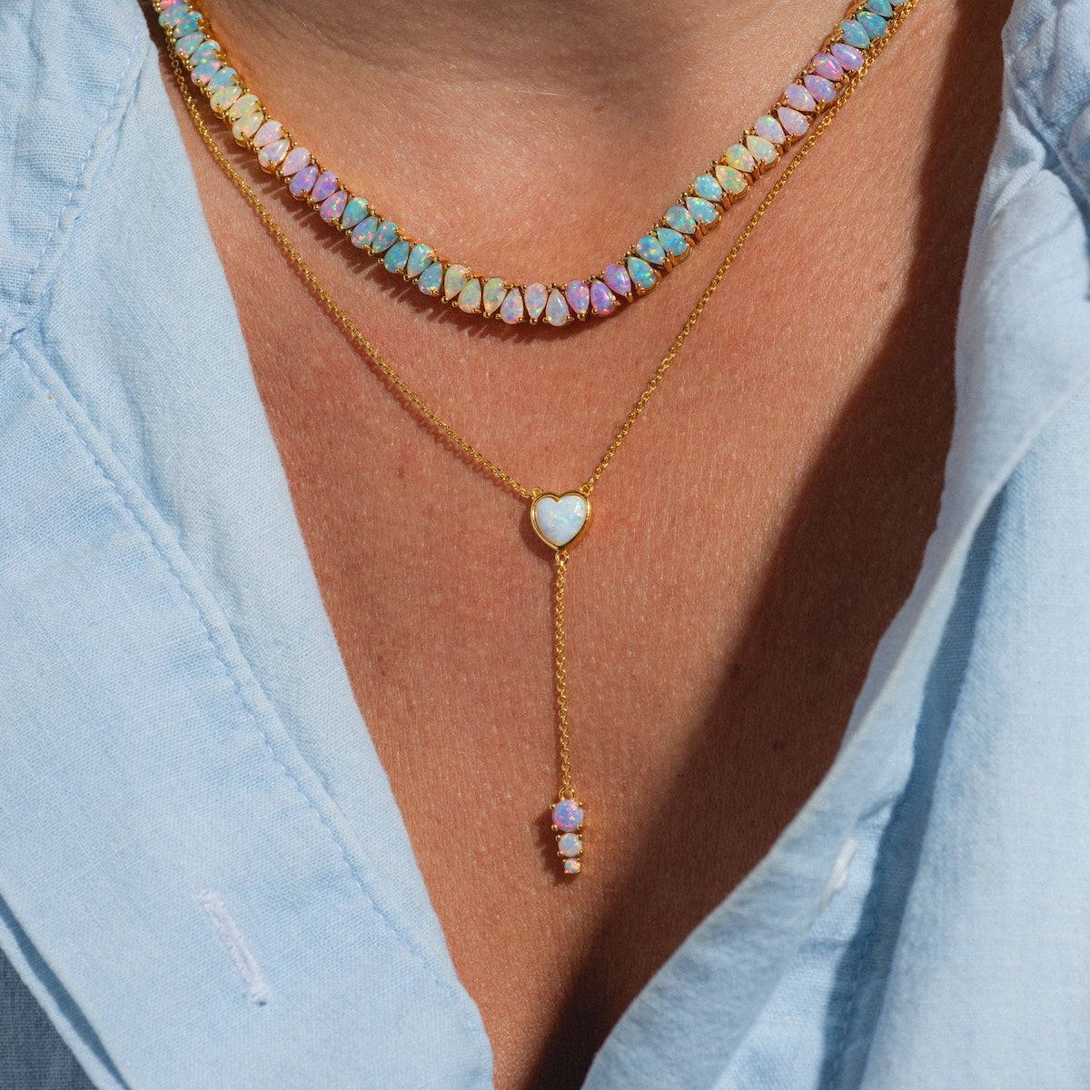 A close-up of a person wearing the Opal Heart Lariat in gold vermeil layered over a light blue button-up shirt.