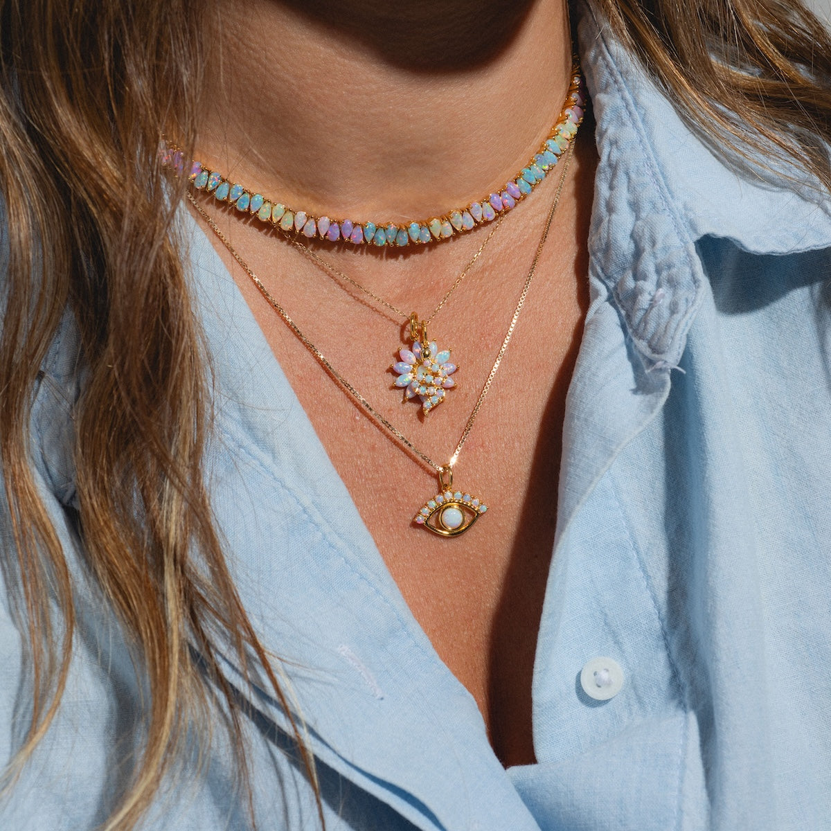 A person wears a light blue collared shirt with layered gold necklaces by Charms by La Kaiser: an 18k gold vermeil floral pendant, a pastel beaded choker, and an eye-shaped pendant featuring a lab-created opal charm.