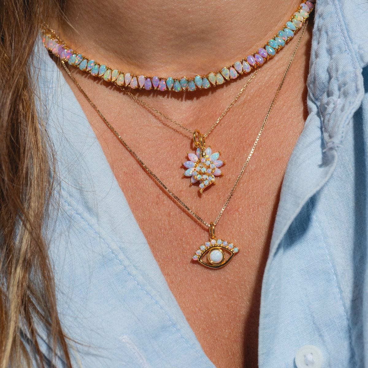 A person wears Charms by La Kaiser: three layered 18k gold vermeil necklaces—a choker with lab-created opal charms, a floral pendant, and an eye pendant—for an elegant, modern touch over a light blue shirt.