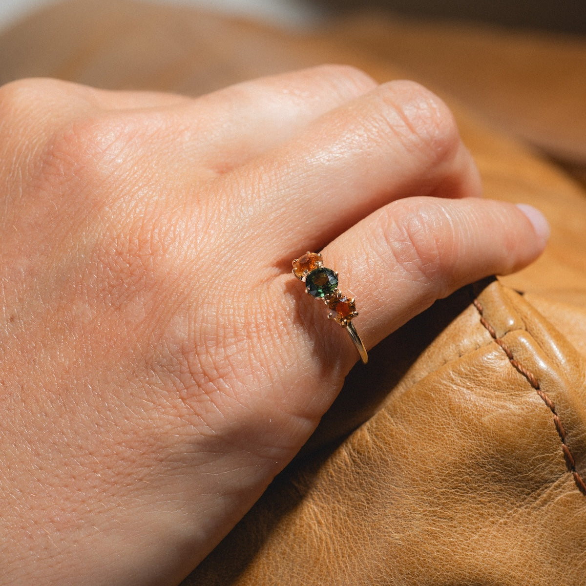 A hand displays the Vintage 14k 1900s Tourmaline Trio Ring in size 4.5, featuring three vibrant gemstones, set in yellow gold and resting on tan leather under natural light.