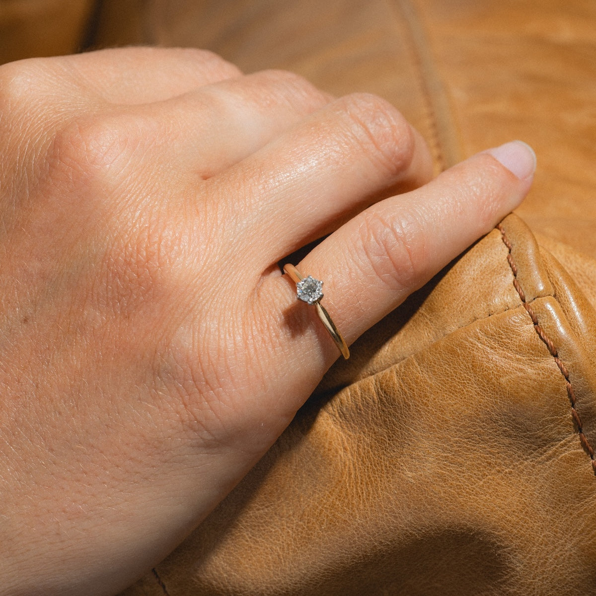 A hand wearing the Vintage 14k 1960s Solitaire Round Diamond Engagement Ring, size 5.5, rests on a light brown leather surface.
