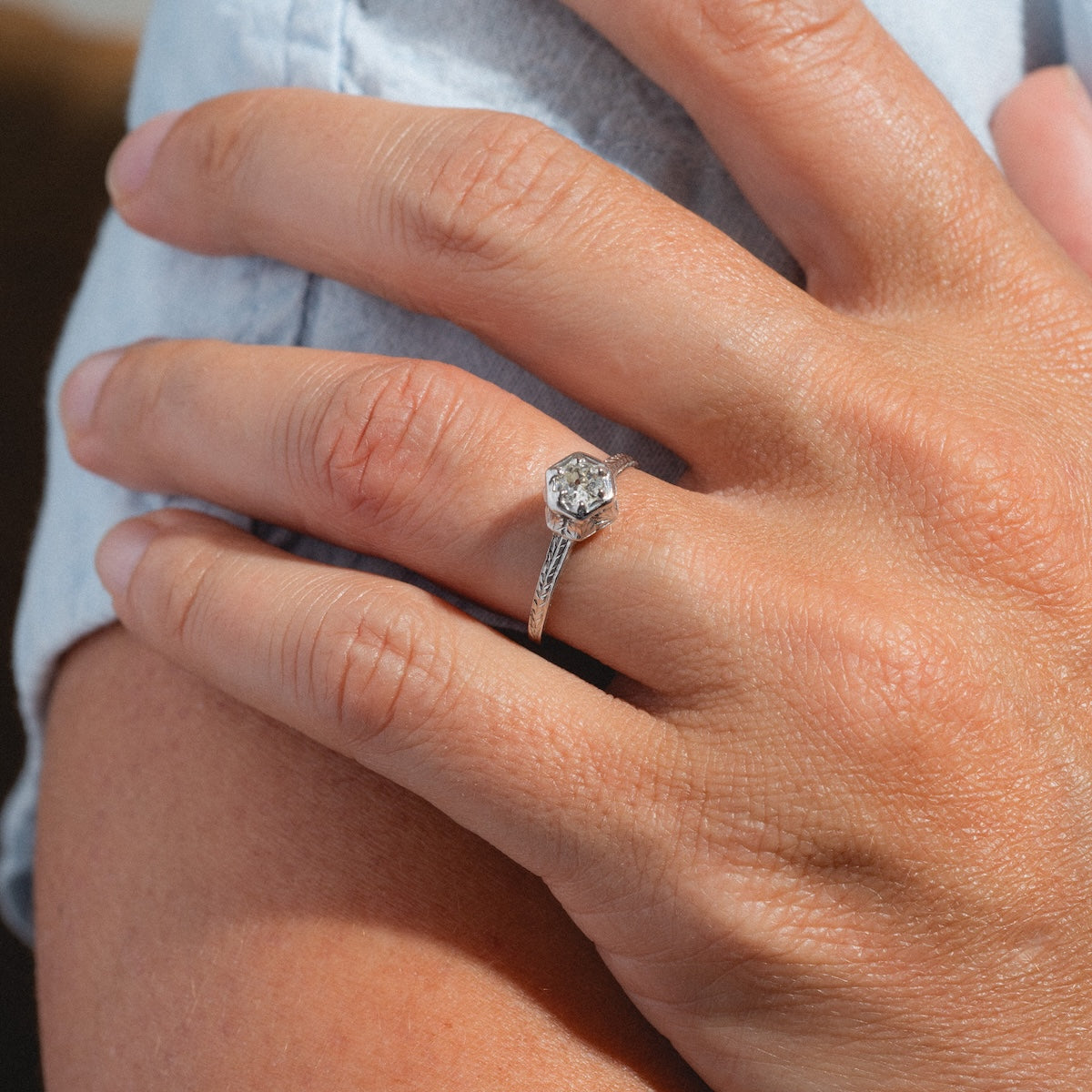 A close-up of a hand wearing the Vintage 18k White Gold Hexagon Set Diamond Ring, size 7, featuring a large round diamond in a hexagonal setting on the ring finger. The hand rests gently on another’s arm; both have light skin tones.