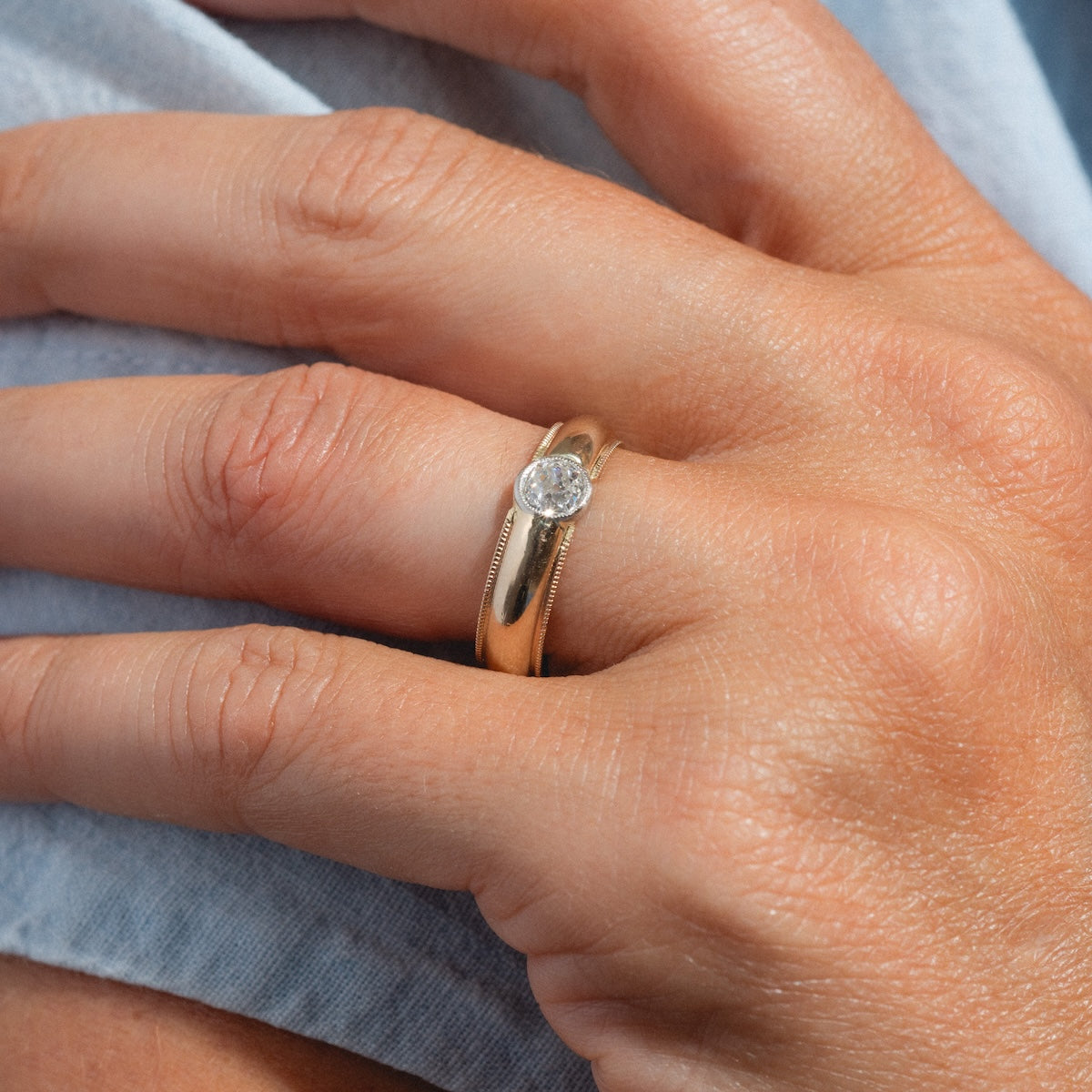 A close-up of a hand wearing two gold rings, including the Vintage 14k 1900s Euro Cut Diamond Classic Band (size 7.5), resting gently on light blue fabric.