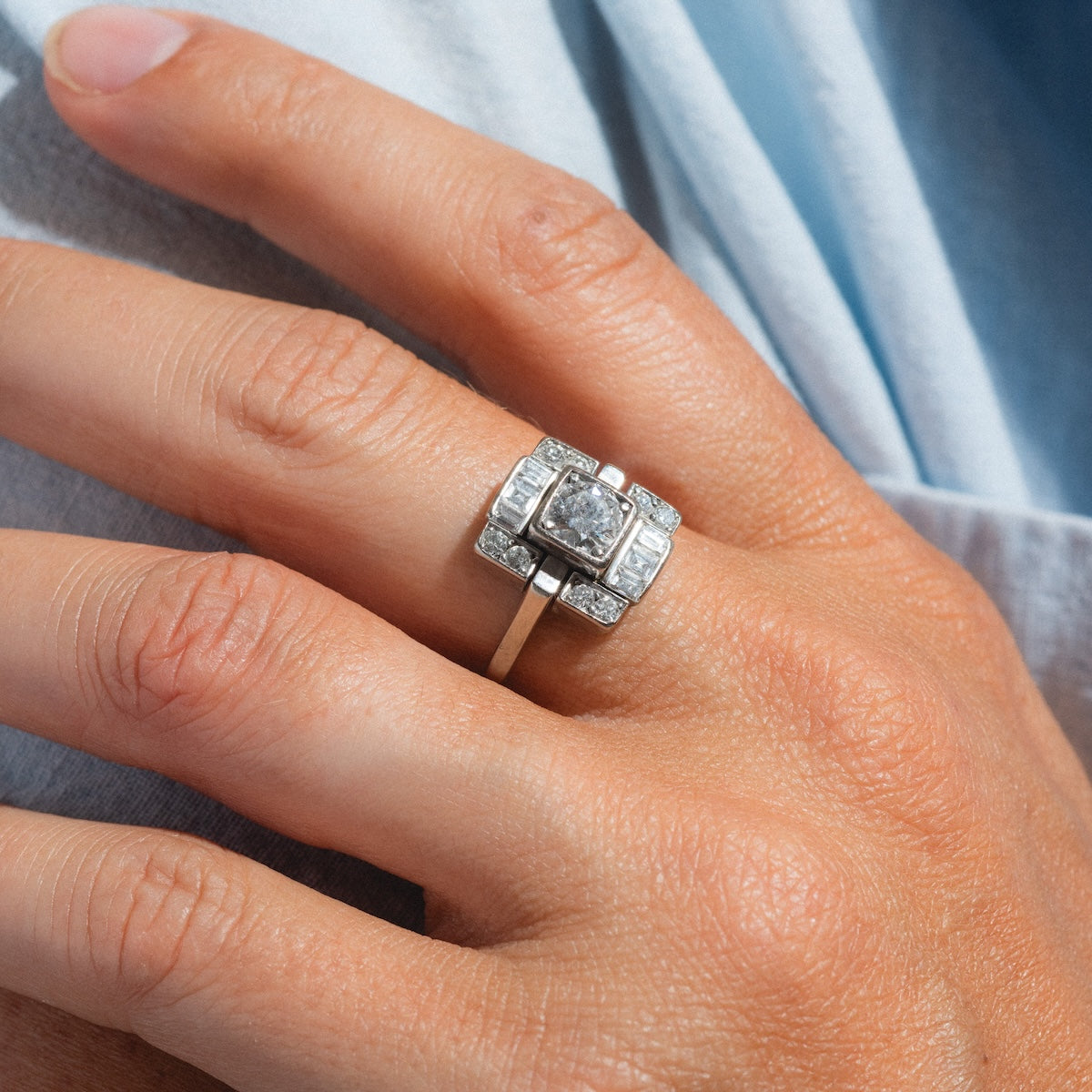 A close-up of a hand wearing the Vintage 18k 1930s Art Deco Geometric Diamond Ring, size 9, with a central square diamond and smaller rectangular and round stones set in white gold against a light blue fabric background.