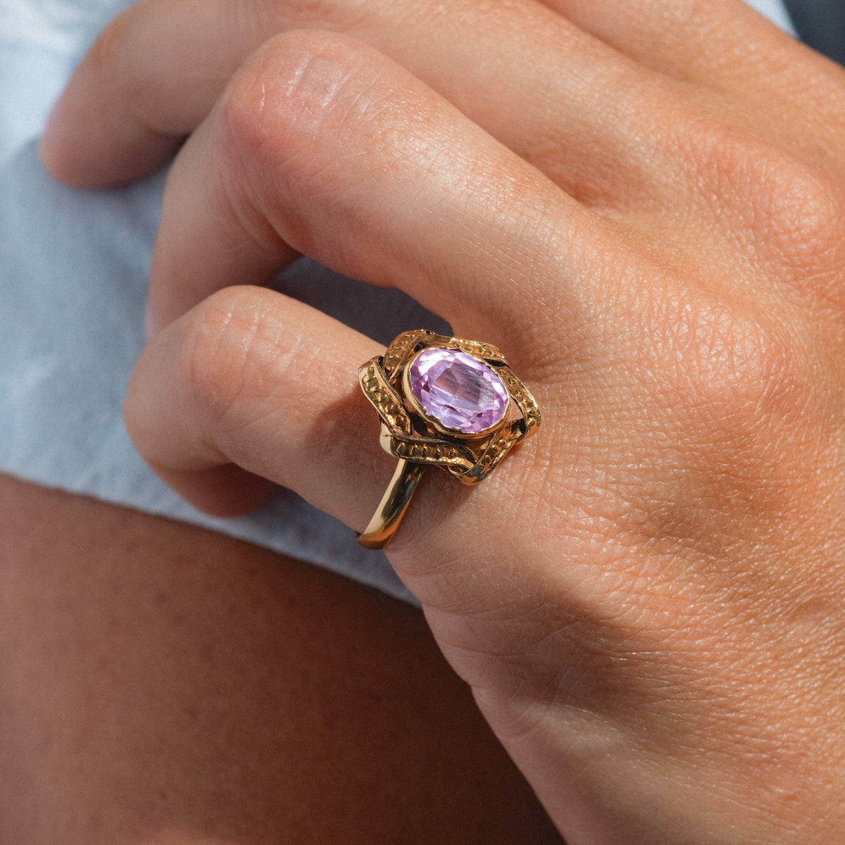 A hand wears the Vintage 18k 1890s Opulent Pink Sapphire Cocktail Ring (Size 6.5), showcasing a large oval sapphire in an ornate band against light blue fabric, capturing Gilded Age elegance.