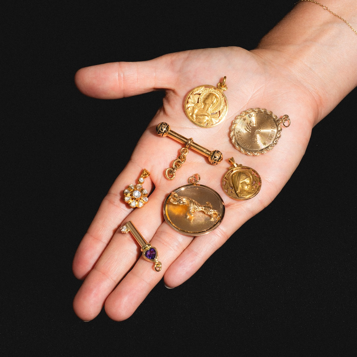 A hand displays several gold pendants against a black background, including an Art Nouveau-inspired Vintage 18k Openwork Mother Mary Coin Charm, two bar-shaped charms, and a gemstone-accented flower charm.