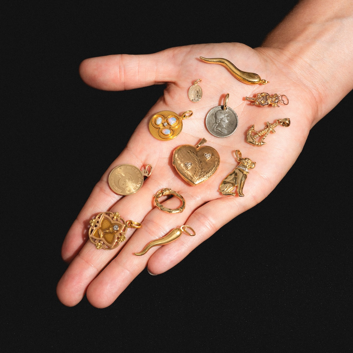 A hand displays several gold and silver pendants—including the Vintage 14k Anchor Charm, hearts, coins, a star, horns, and other intricate designs—showcasing unique jewelry against a black background.