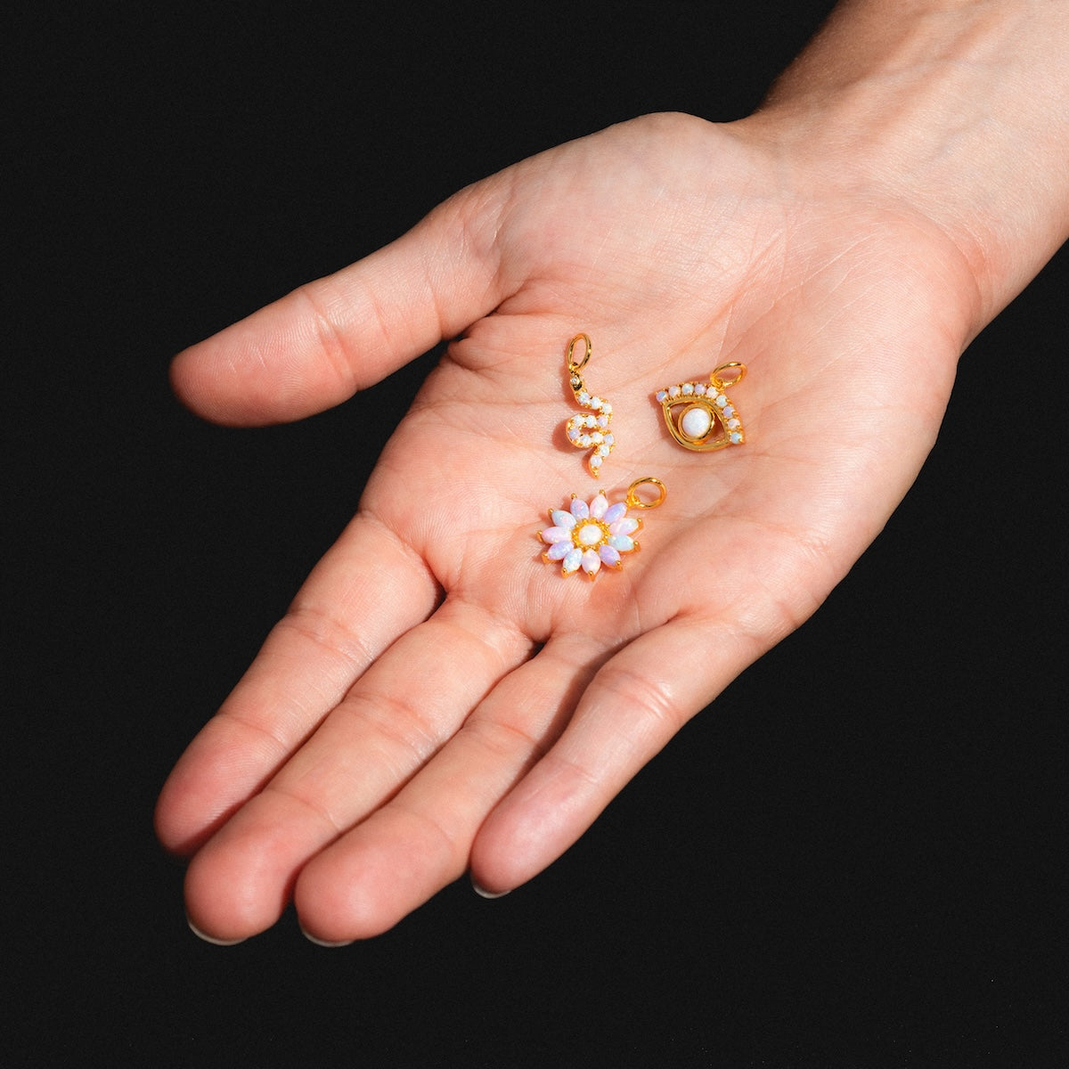 A hand holds three Charms by La Kaiser—an 18k gold vermeil daisy, a sparkling snake, and an eye with a central pearl—set against a black background.