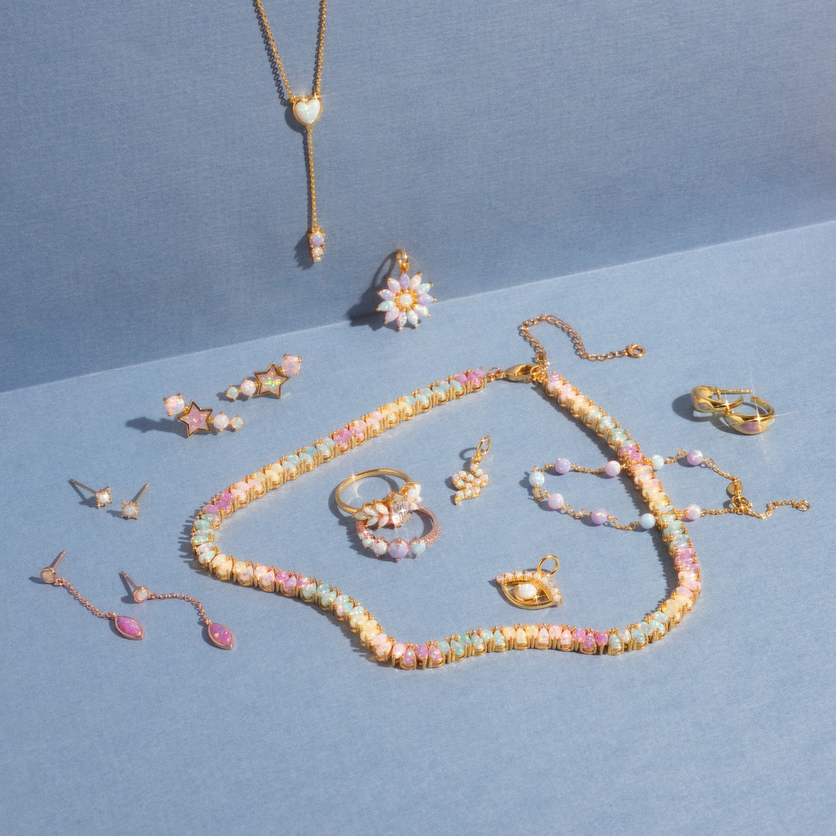 A selection of gold and pastel jewelry, including the Opal Heart Lariat necklace, rings, earrings, and gold vermeil bracelets displayed on a blue surface under gentle lighting.
