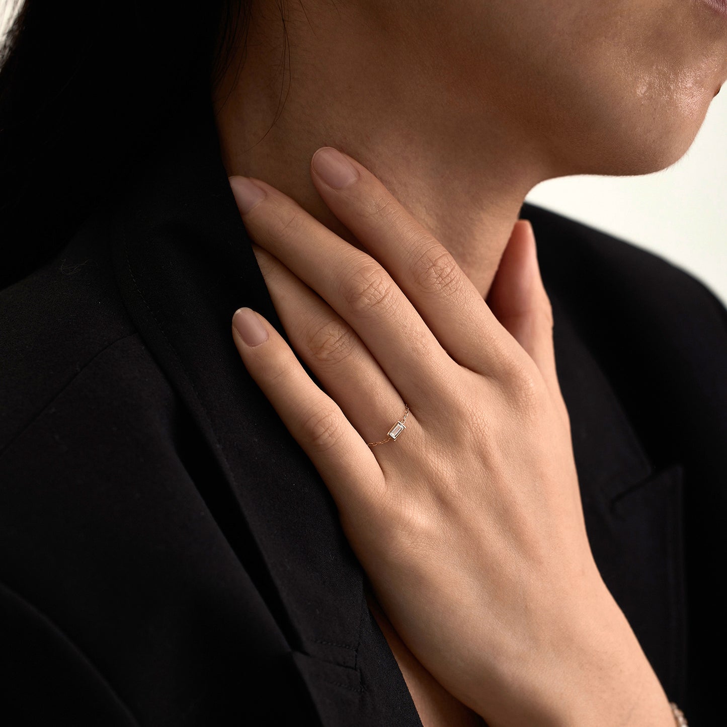 A person in a black blazer touches their neck, showcasing the Solid Gold White Topaz Baguette Chain Ring on their finger. Their short nails are neatly manicured with a natural finish.