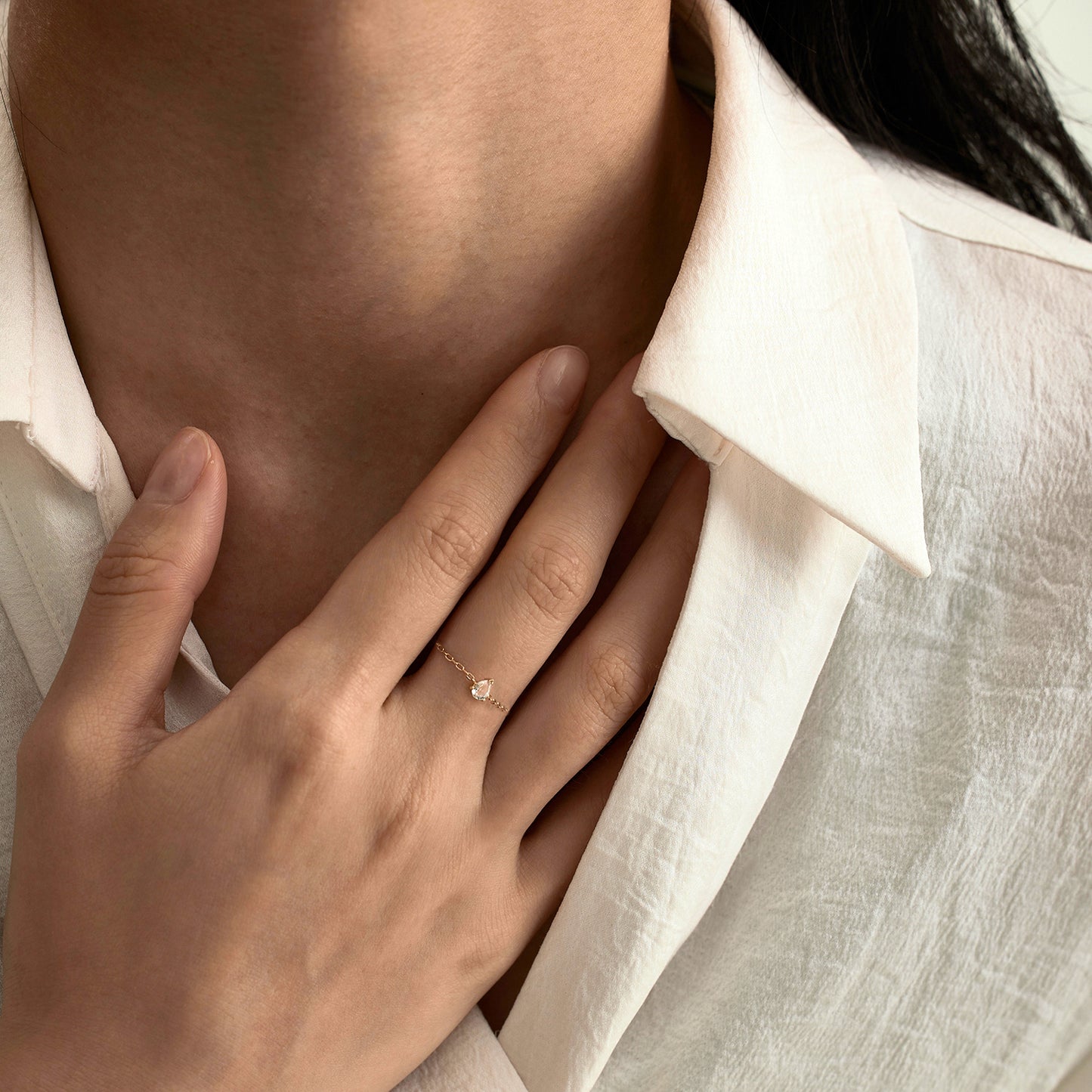 A person in a textured white collared shirt touches their neck, showcasing the Solid Gold White Topaz Pear Chain Ring on their finger as its delicate shimmer catches the light.