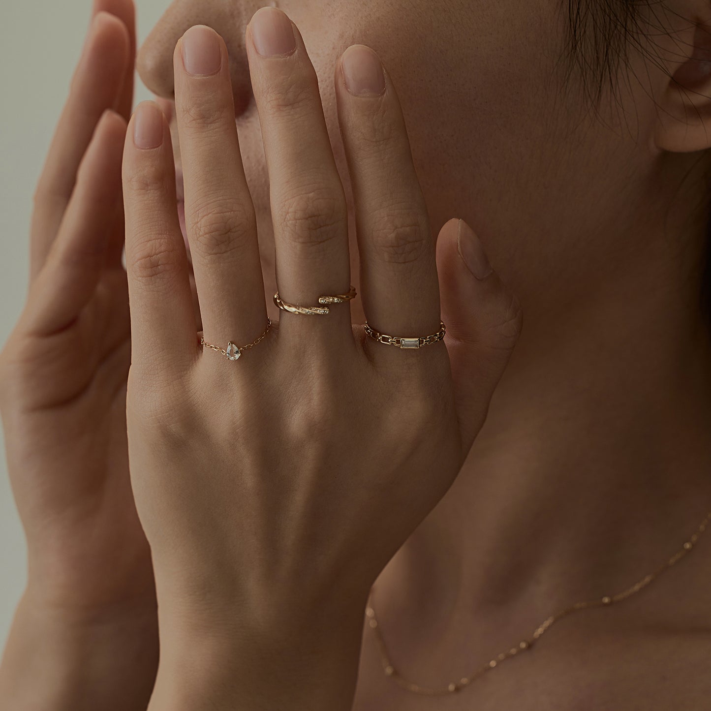 A person with manicured nails covers part of their face with both hands, styled in a Solid Gold White Topaz Pear Chain Ring, another gold band, and a thin gold necklace. A softly blurred background adds elegance.