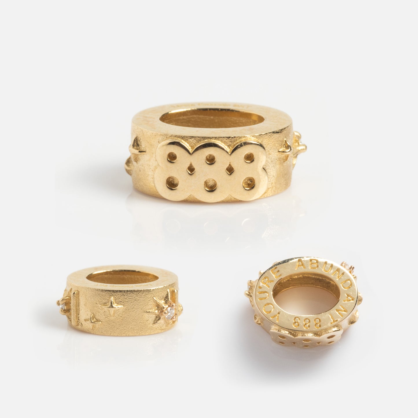 Three textured gold beads with engraved patterns and star-shaped accents rest on a plain white background. One bead, the Solid Gold Angel Number Spacer Charm—a Local Eclectic Exclusive—features an inscription along its edge.