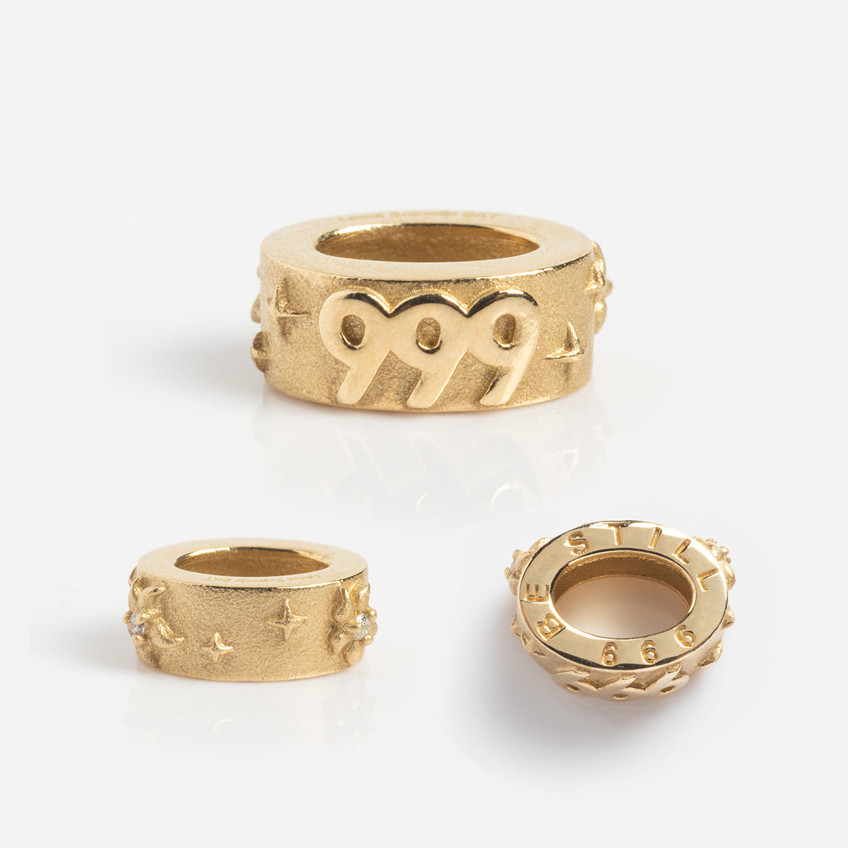 Three gold rings are displayed: one is a Local Eclectic Exclusive with 999 and heart designs, another has star and cross patterns, and the third is engraved with STILL and 666. All feature the Solid Gold Angel Number Spacer Charm.
