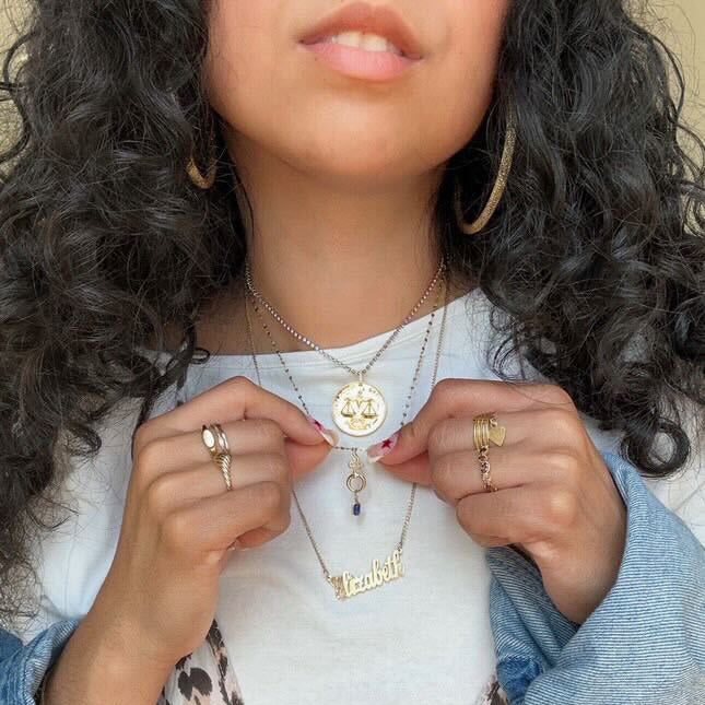 A woman with curly hair wears multiple gold necklaces, including a Solid Gold Charm Clip, personalized pieces, and a circular pendant. She displays the necklaces with rings on her fingers, hoop earrings, a white top, and a denim jacket.
