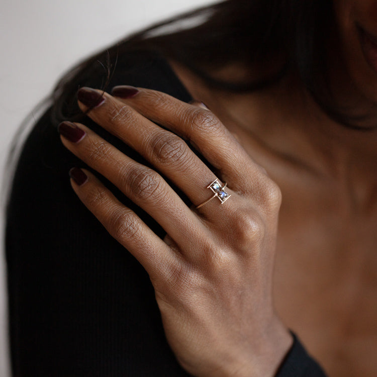 A womans hand with dark polished nails wears the Amulet Hourglass Ring, a rectangular moonstone piece set in 14k solid yellow gold, perfectly complementing her black long-sleeved top.