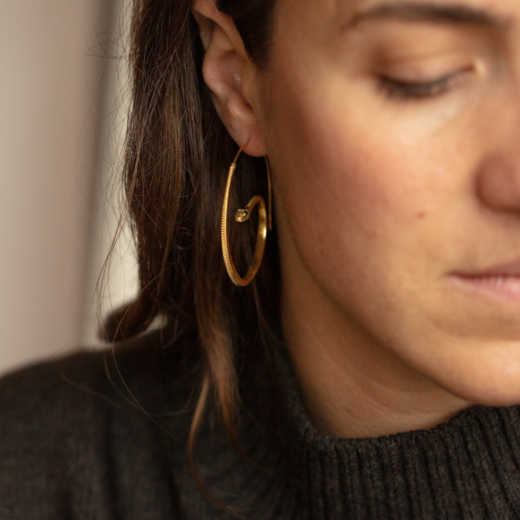 A close-up shows a woman with brown hair wearing a dark knit sweater and Snake Hoop Earrings in gold. The softly lit image focuses on her ear, highlighting the bold snake-shaped hoops, with only part of her face visible.