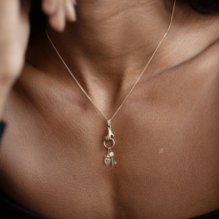 A close-up shows a person with dark skin wearing Acanthus 14k Solid Gold Tiny Charms—a claw, heart, and key—on a gold necklace. Their black top sits slightly off the shoulder.