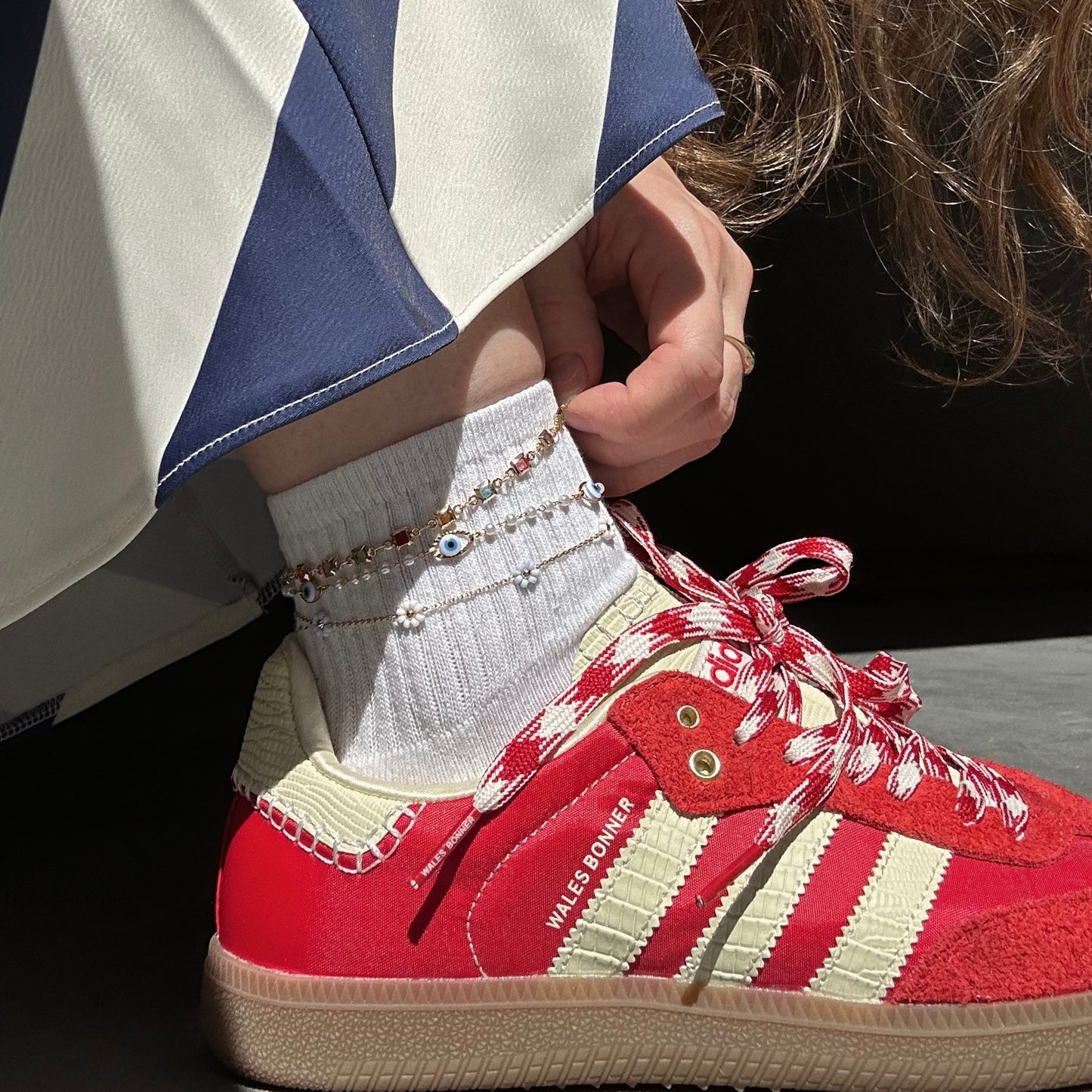 A person wearing red and white Adidas sneakers, white socks, blue-and-white pants, and a gold plated Evil Eye Anklet featuring a delicate evil eye charm is shown.