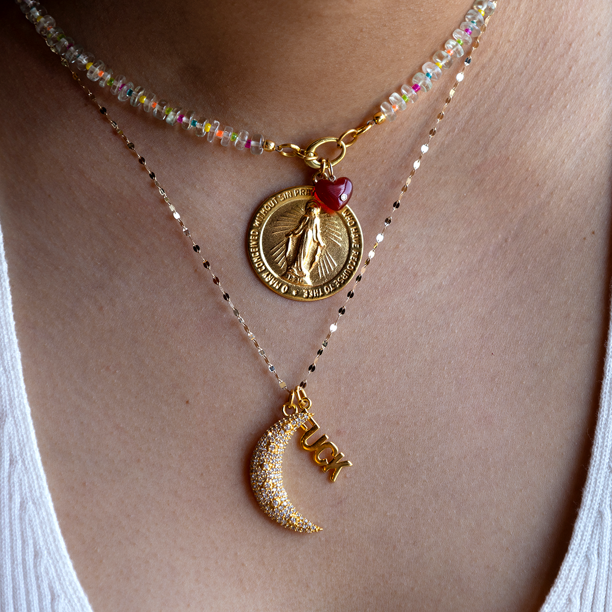 Close-up of a person in a white top wearing Charms by Anokhi Green: three layered necklaces including a beaded choker, gold coin pendant with red bead, and gold chain with cubic zirconia charms spelling “LUCK.”.
