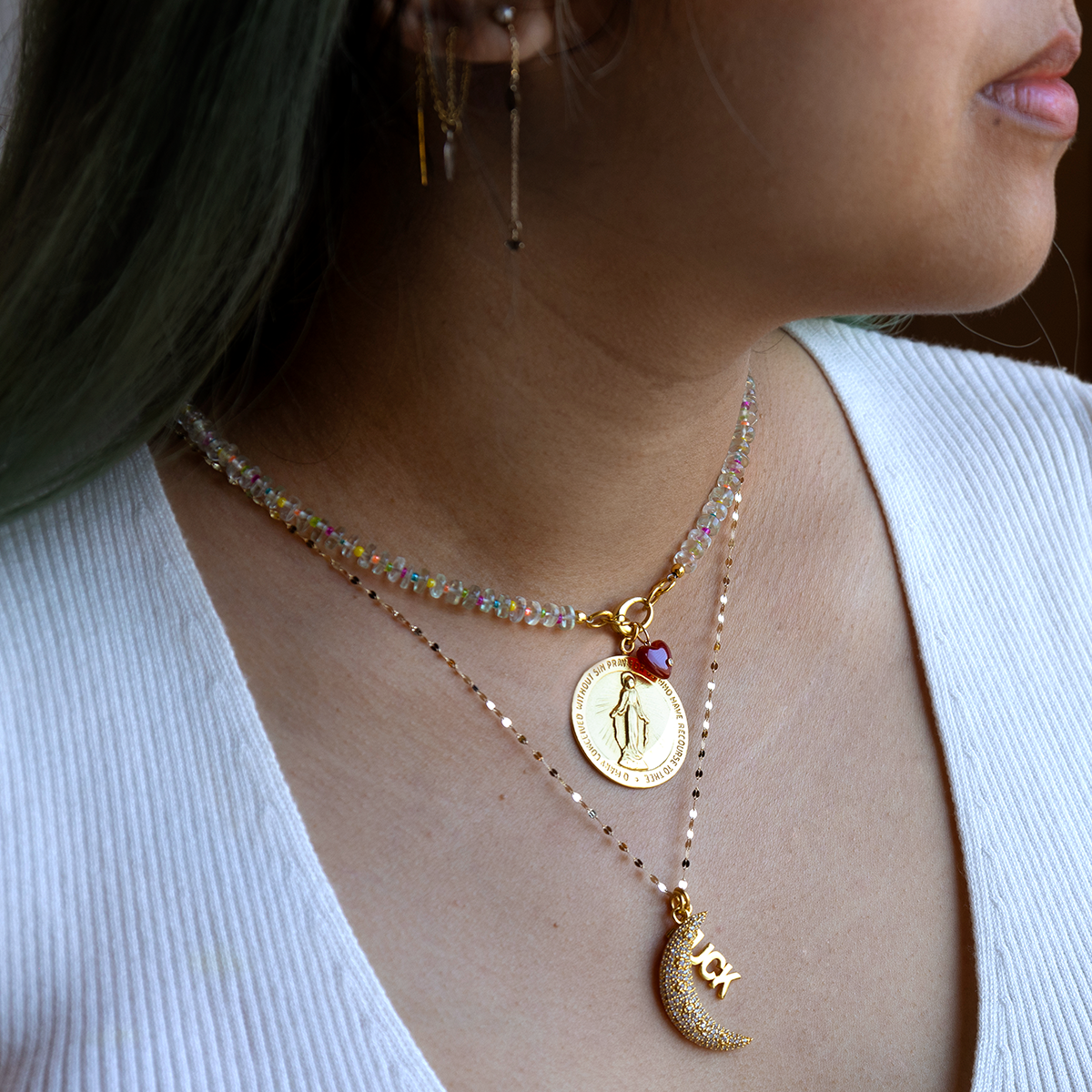 A woman wears Charms by Anokhi Green: a white ribbed top, two layered gold-plated necklaces—one with a coin and red heart charm, the other with an inscribed crescent moon—and two gold earrings.