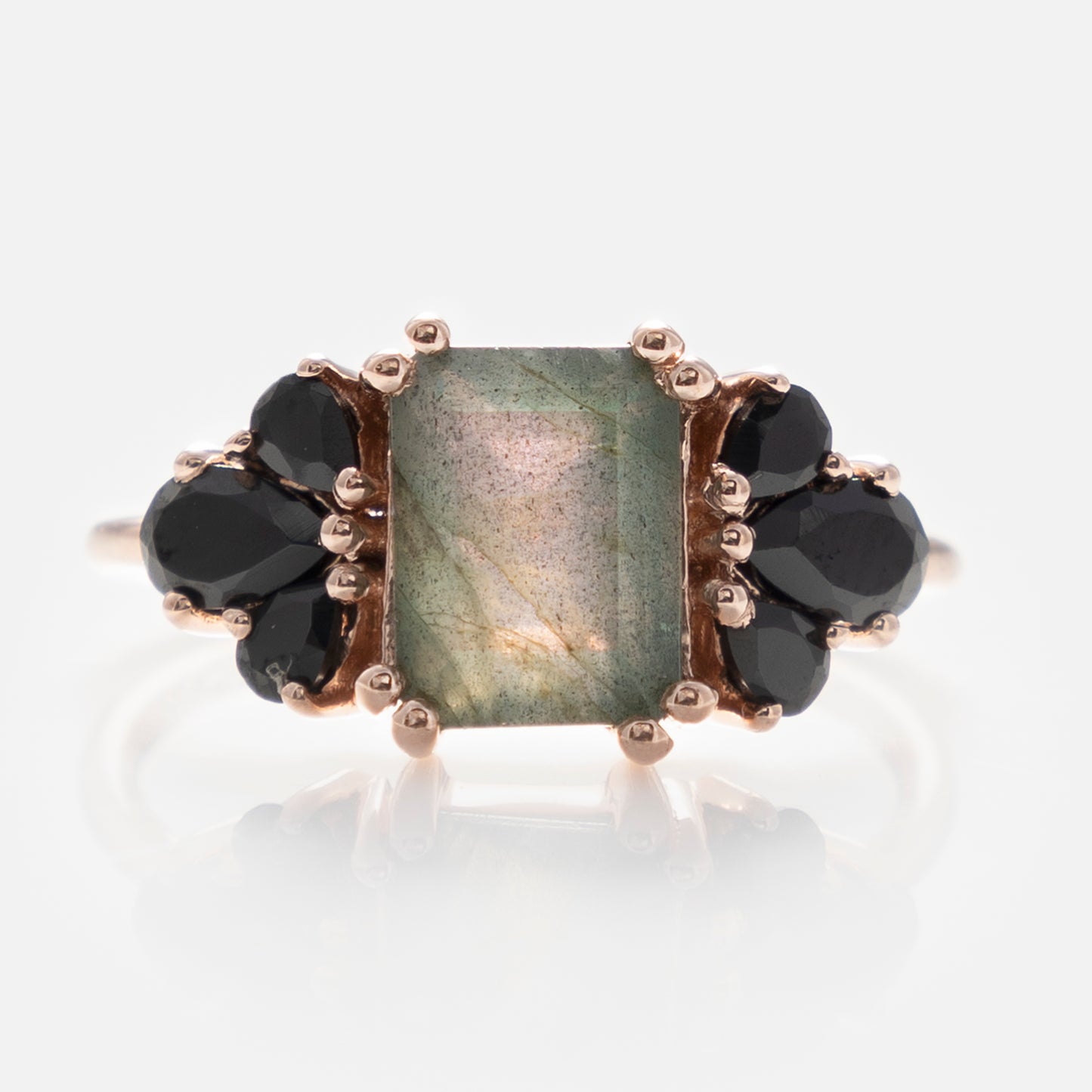 The Gemstone Empress Ring is a gold statement piece with a rectangular iridescent green labradorite center, accented by clusters of round and pear-shaped black stones on each side.
