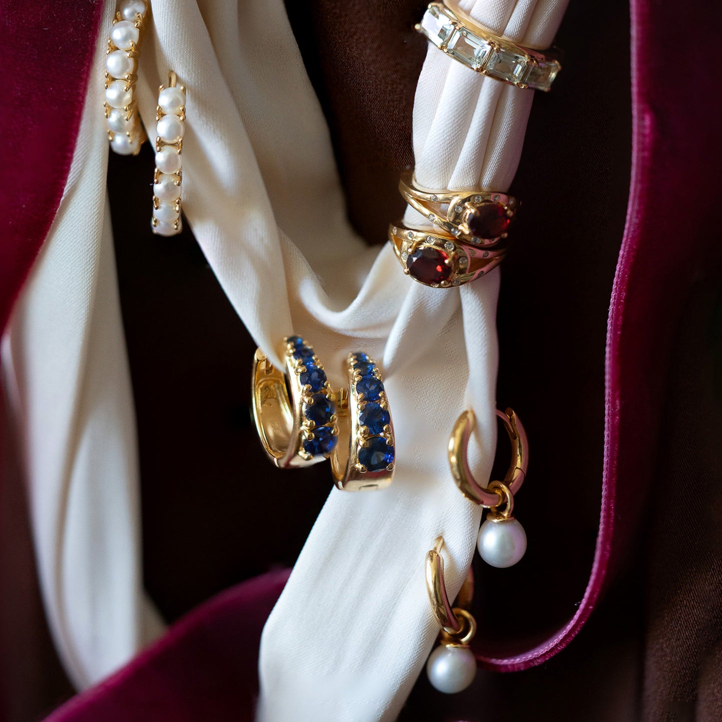 A close-up of vintage-inspired jewelry, including 14k gold vermeil rings, pearl earrings, and Sapphire Soliloquy Huggie Hoops, displayed on white fabric with burgundy velvet.